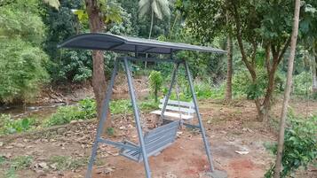A metal swing set with a bench, surrounded by greenery and a small stream.