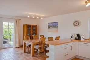 A dining area with a wooden table and chairs, a cabinet with glass doors, a painting of a beach scene, a clock on the wall, and a kitchen area with a coffee maker. - Holiday home with 3 bedrooms (Heringsdorf)