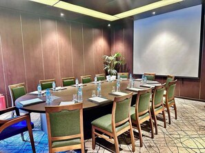 Un salón de conferencias con una gran mesa, sillas, botellas de agua y una pantalla para proyector.