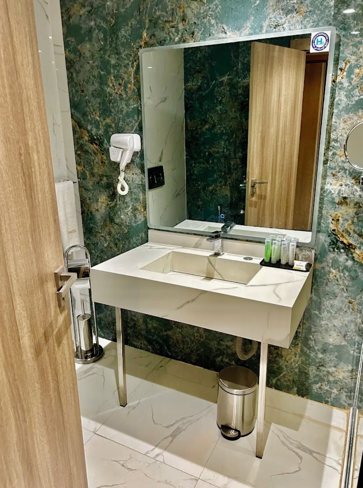 Un baño con pared de mármol, lavabo doble, espejo, secador de pelo y cubo de basura.