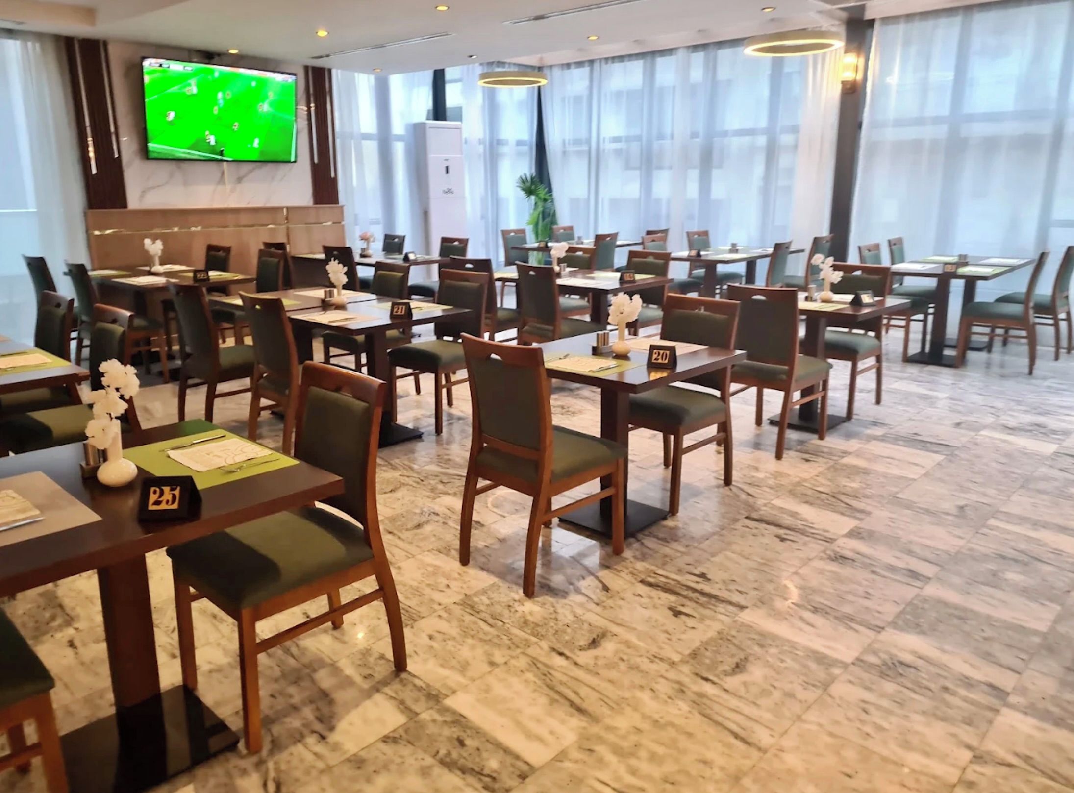 Comedor de hotel con varias mesas y sillas, un televisor en la pared y amplias ventanas.