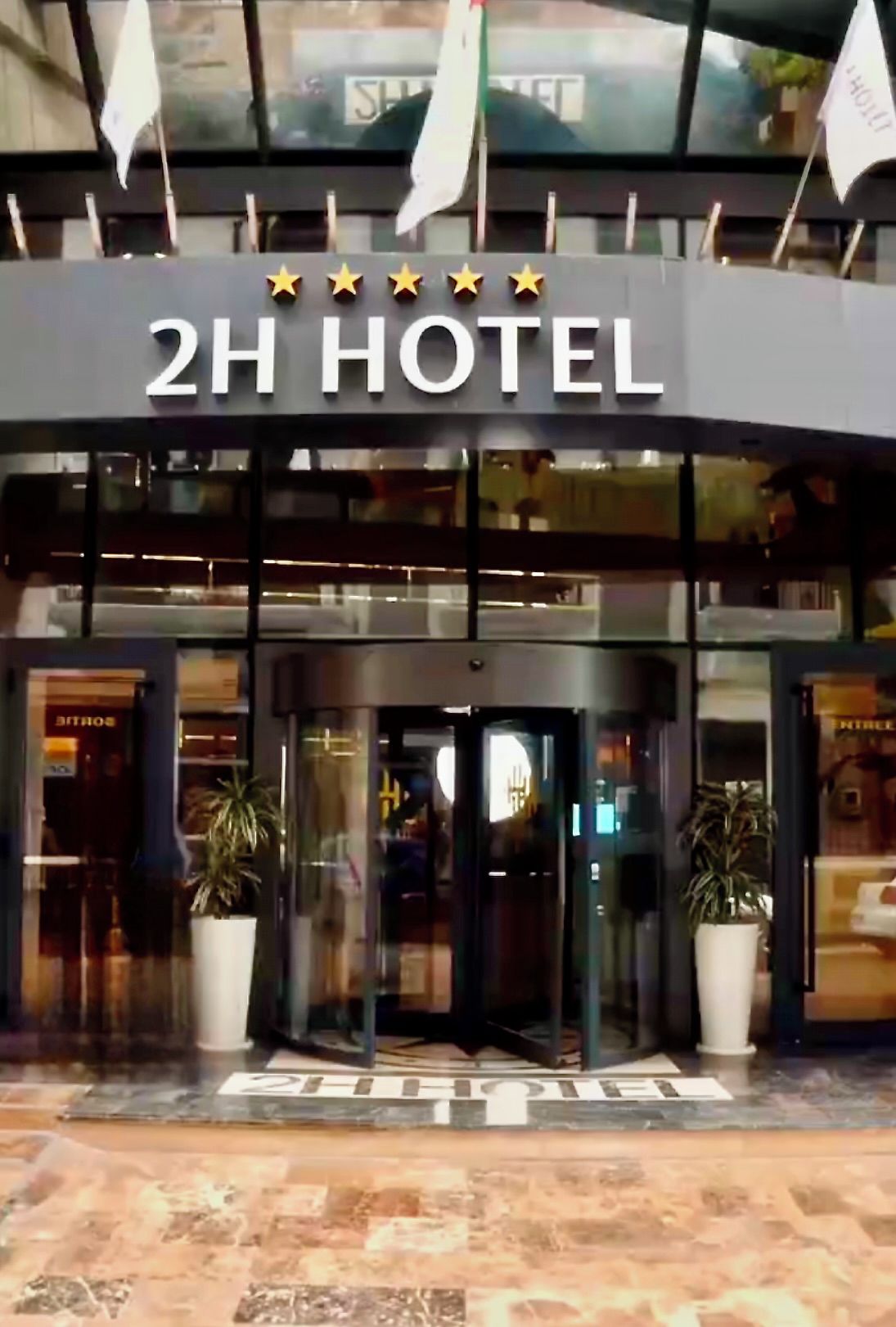 La entrada a un Hotel 2H con fachada de vidrio, plantas en macetas y un cartel con la clasificación de estrellas.