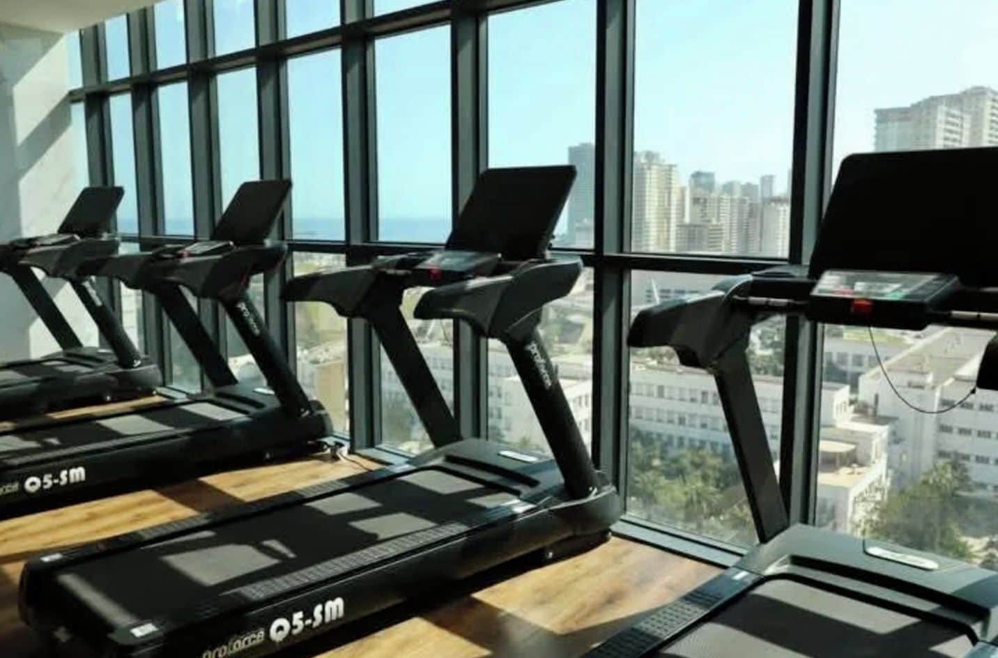 Un gimnasio con cintas de correr alineadas frente a amplias ventanas con vista a la ciudad.