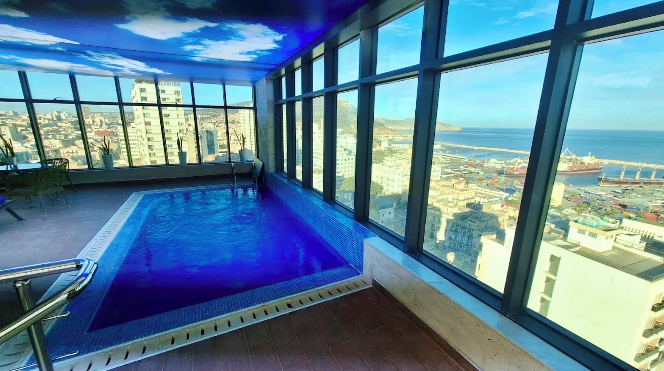 Une piscine intérieure avec vue sur la ville, de grandes fenêtres et un plafond décoré de motifs nuageux.