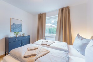 A hotel room with two beds, a bedside table, a window with curtains, and a wall-mounted artwork. - Hi there, and welcome to the Küstenkoje! (Dangast)