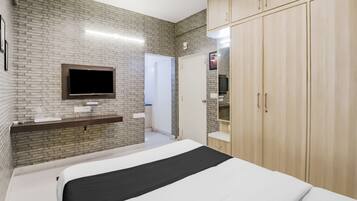 A bedroom with a brick wall, a flat-screen TV mounted above a vanity, a bed with a black and white bedspread, and wooden cabinets.