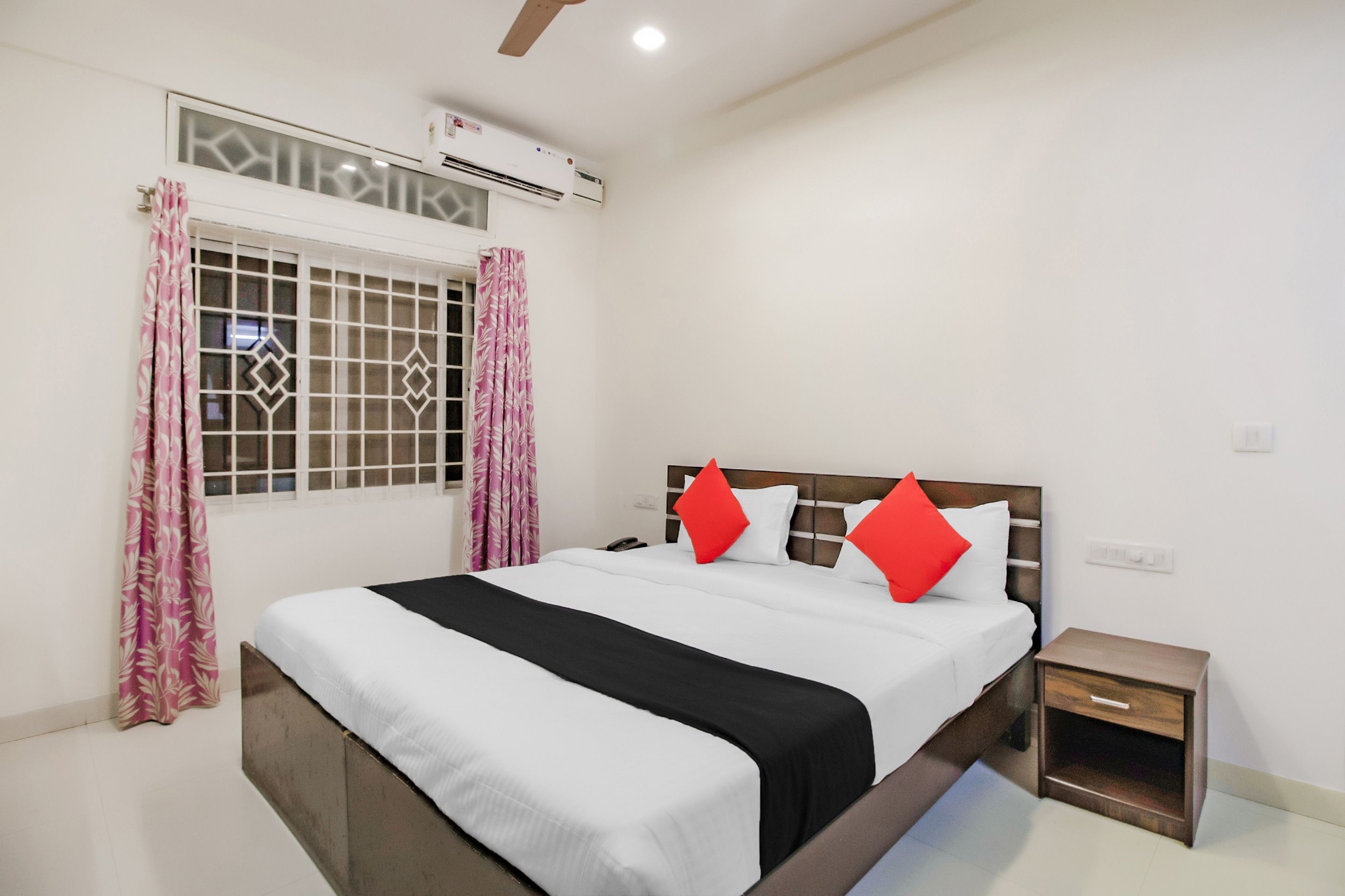 A hotel room with a bed, a bedside table, a nightstand, a window with curtains, and an air conditioning unit.