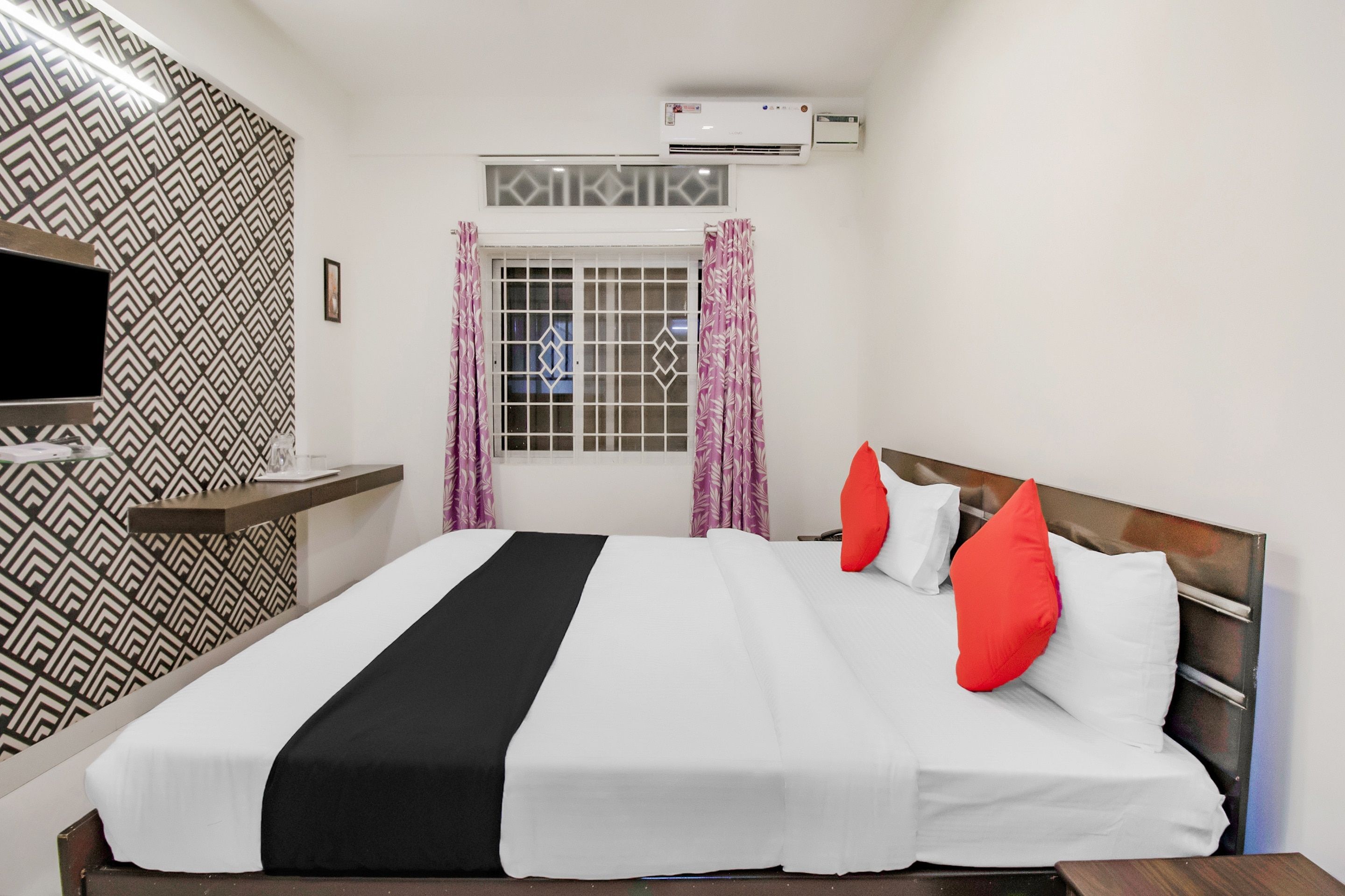 A hotel room with a bed, a TV, an air conditioning unit, and a window with curtains.