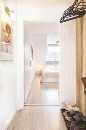 A hallway leading to a bedroom with a bed, a nightstand, and a window. There is a shoe rack with various slippers and a wall-mounted rack with items. - Vrbo Property (Meerbusch)