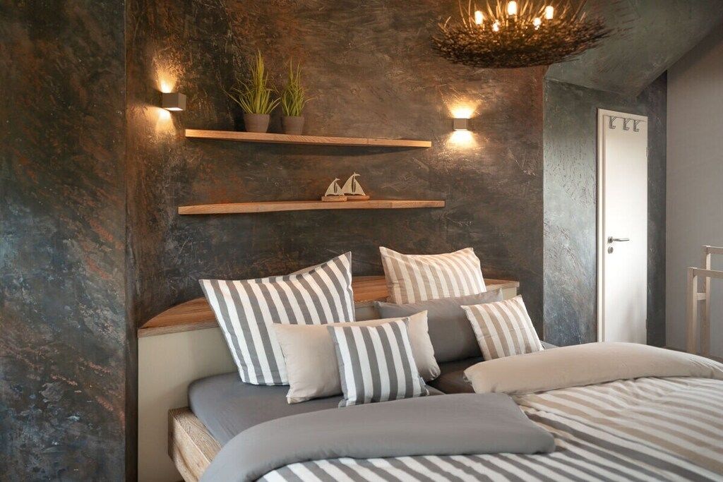 A bedroom with a bed, striped pillows, a wall-mounted shelf with plants, and a door with a number.