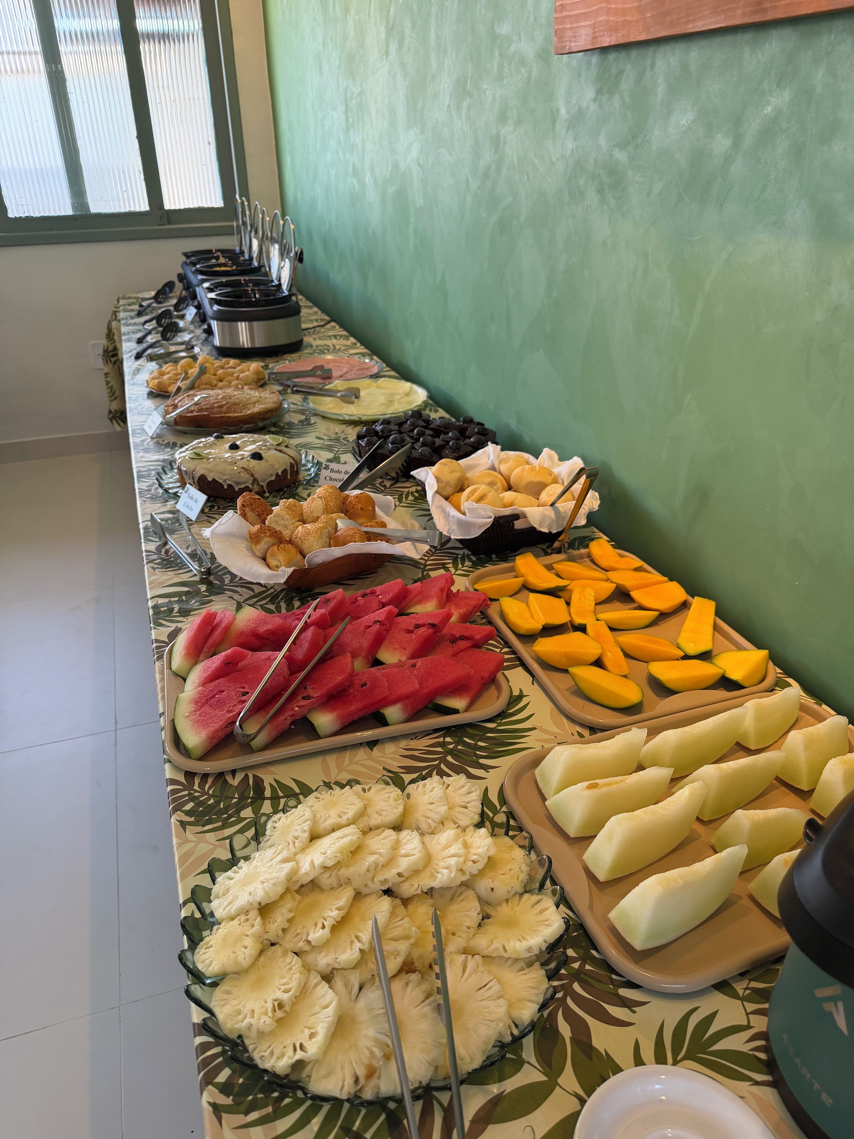 Mesa de buffet com diversas opções de comida, incluindo frutas, doces e uma máquina de café.