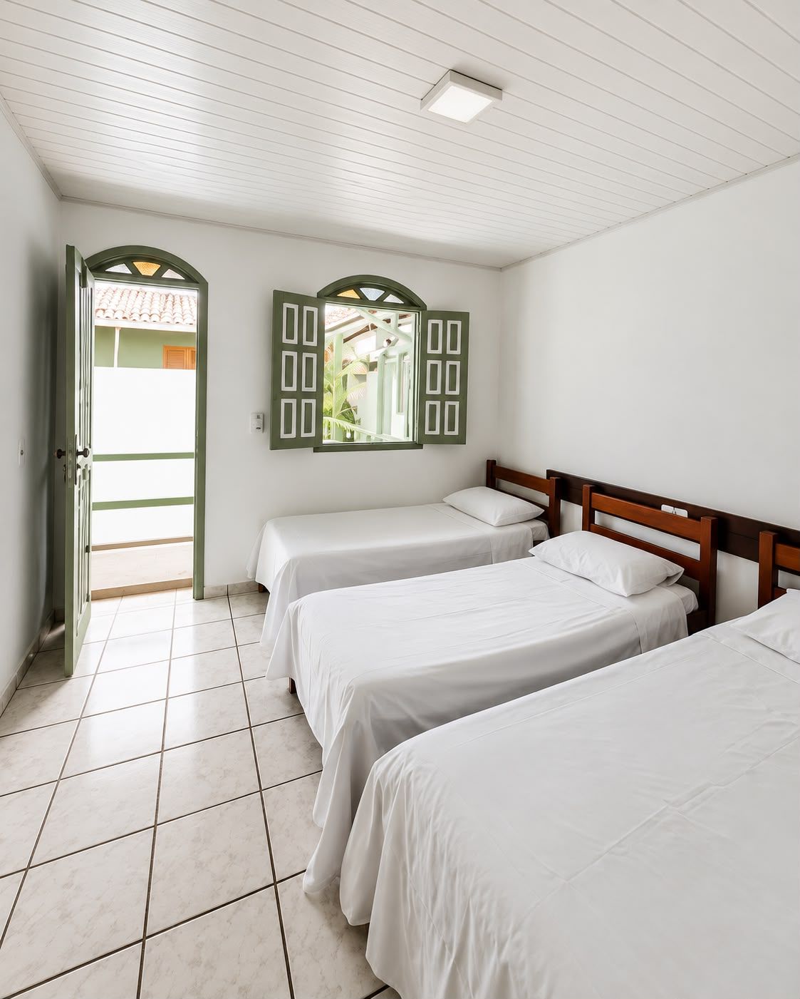 A room with three single beds, a tiled floor, a door leading outside, and a window with shutters.