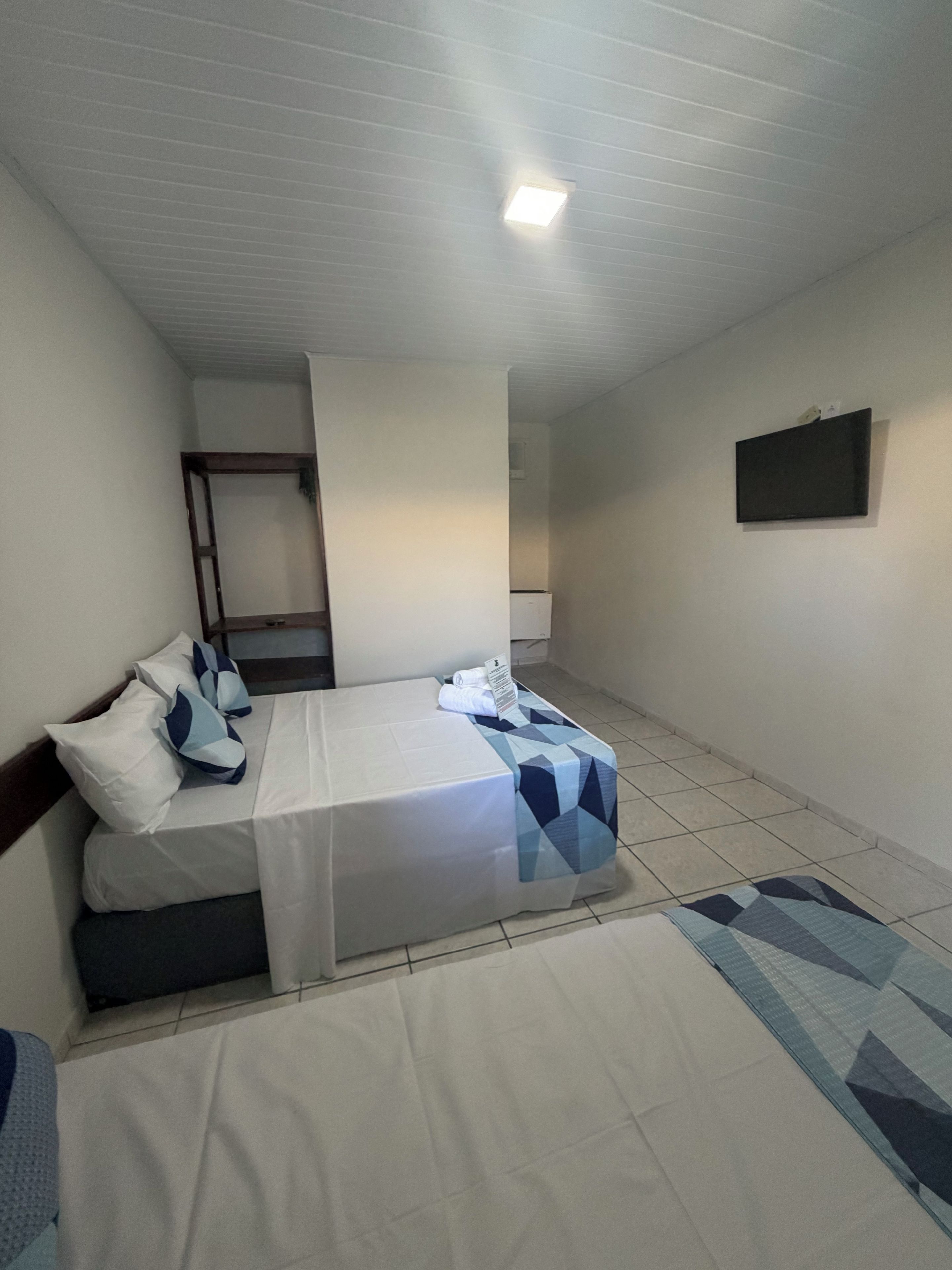 Una habitación con una cama, una mesita pequeña, un televisor en la pared y una lámpara en el techo.
