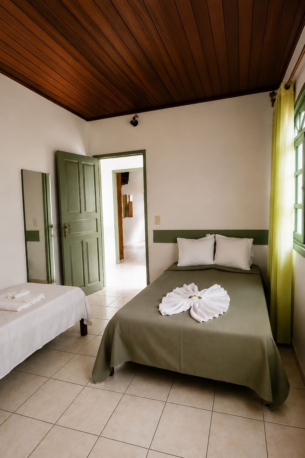 A hotel room with a bed, a smaller bed, a green door, a mirror, and a window with curtains.