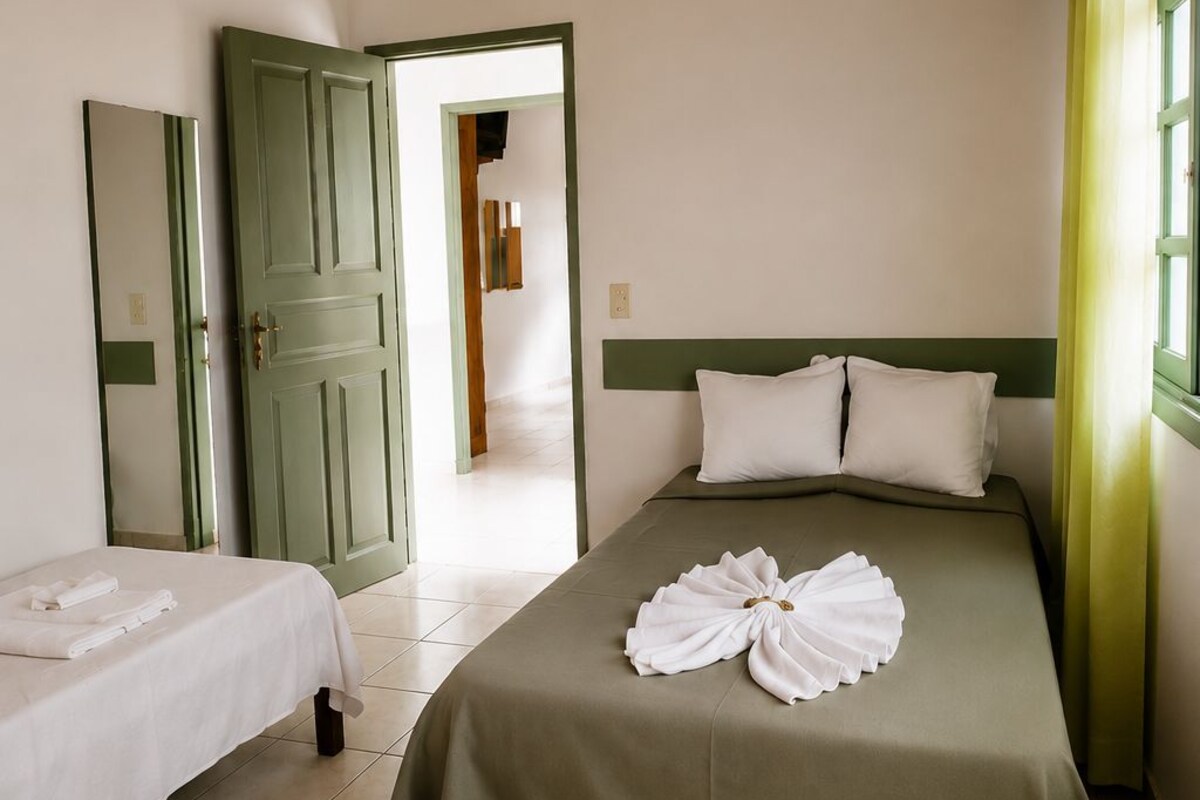 A hotel room with a bed, a smaller bed, a green door, a mirror, and a window with curtains.