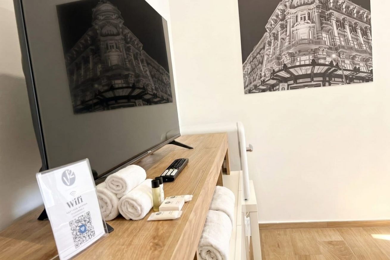 Una camera d'albergo con una scrivania in legno, una TV, il simbolo del WiFi, asciugamani arrotolati e un quadro incorniciato raffigurante un edificio.