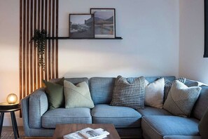 A modern living room with a grey sofa, patterned cushions, a small side table with a lamp, and a wall-mounted shelf with framed pictures. - Cabot Peak Retreat - This professionally curated, design-forward condo is your perfect basecamp, located just minutes from both downtown and the world-class slopes of Revelstoke Mountain Resort. (Revelstoke)