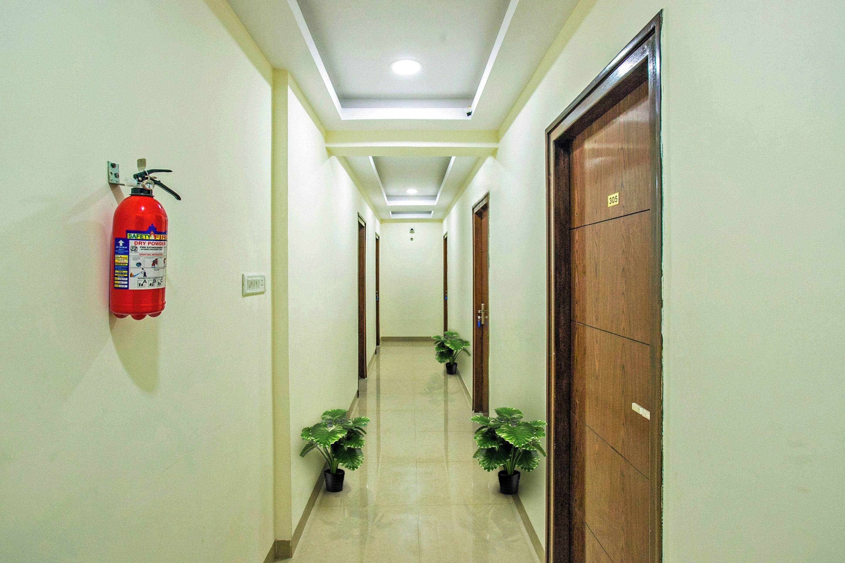 A hallway with a fire extinguisher mounted on the wall, potted plants, and a wooden door with a number.