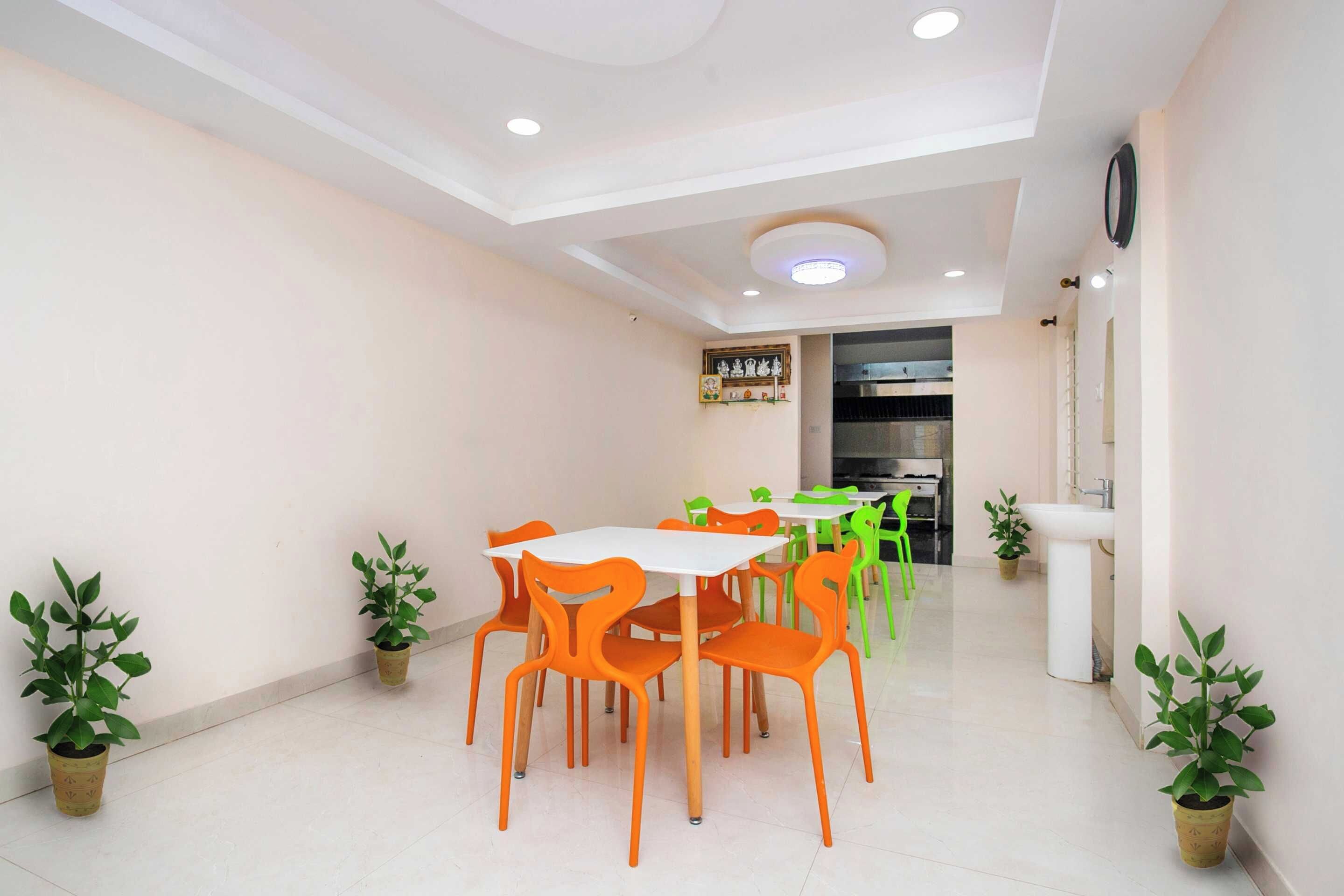 A dining area with a white table and orange chairs, potted plants, and a kitchen in the background.