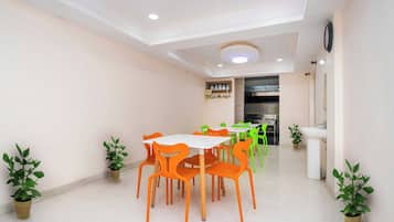 A dining area with a white table and orange chairs, potted plants, and a kitchen in the background.