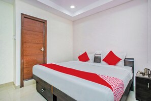 Una habitación de hotel con una cama grande, una puerta de madera, una mesita de noche con una lámpara y un mueble pequeño.