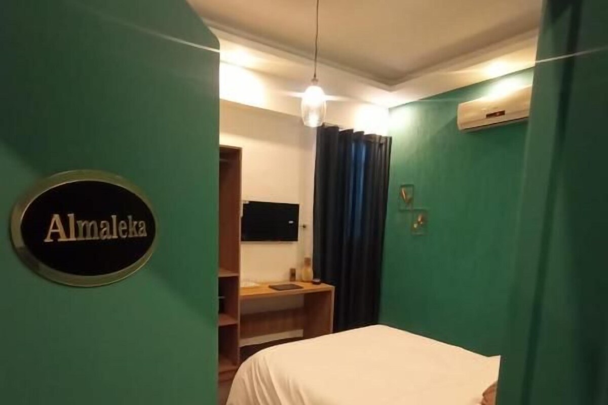 A room with a bed, a sink, and a mirror. There is a sign on the wall that reads 'Almaleka.'