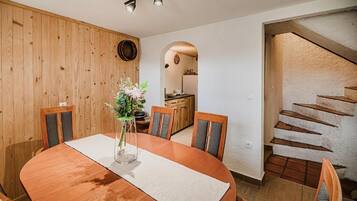 A dining area with a wooden table and chairs, a vase with flowers, and a kitchen visible through an archway.