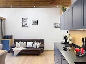En moderne stue med en mørkebrun sofa, træloft og et køkkenområde med grå skabe og en skål med frugt.