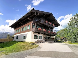 A two-story chalet with a balcony, a wooden deck, and a mountain backdrop.