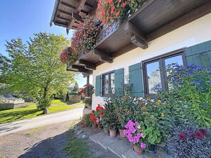 Una casa con balcón decorado con flores, camino de grava y jardín con diversas plantas.