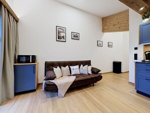 A modern living room with a brown sofa, a television on a wooden stand, and a kitchen area with cabinets and a microwave.