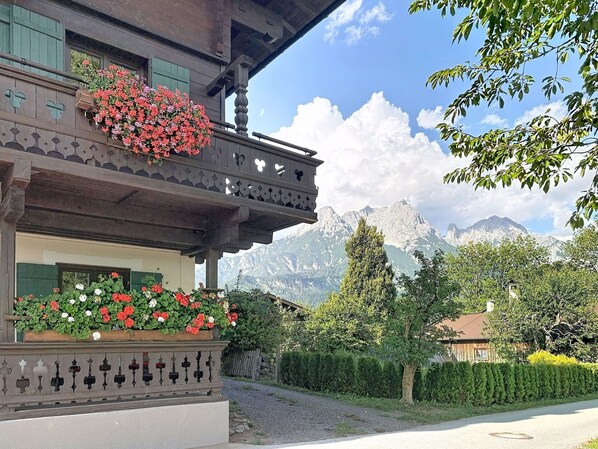A traditional chalet with a balcony adorned with flowers, set against a backdrop of mountains.