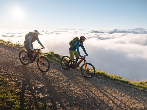 Due ciclisti su un sentiero di montagna con vista su nuvole e montagne in lontananza.