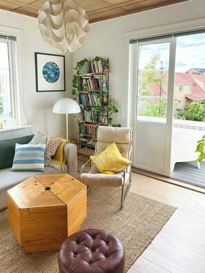 A living room with a sofa, a chair with a yellow pillow, a wooden coffee table, a bookshelf, and a large window with a view of buildings. - Flat with garden in historic house beside Sagene (Oslo)