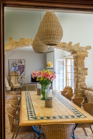 A dining area with a large table, wicker chairs, a hanging light fixture, and a stone wall. - Villa La Roche d’Or | Panoramic Pool (Tourrettes-sur-Loup)