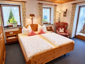Ein Doppelbett mit floraler Bettwäsche, ein roter karierter Sessel, ein Nachttisch mit Lampe und ein Fenster mit Blick auf Grünflächen.