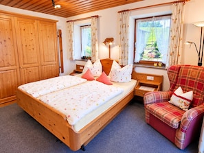 Ein Schlafzimmer mit Doppelbett, Holzschrank, zwei Nachttischen, einem Sessel und einem Fenster mit Vorhängen.