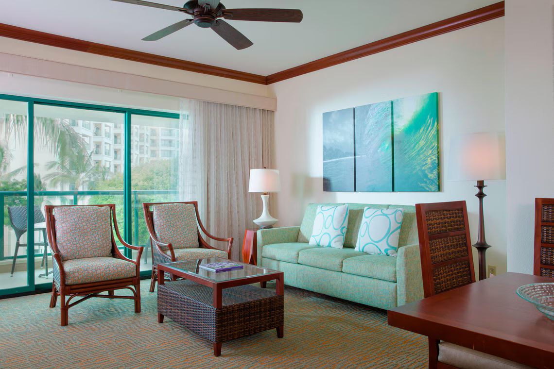 A hotel room with a balcony, a sofa, two armchairs, a coffee table, and a ceiling fan.