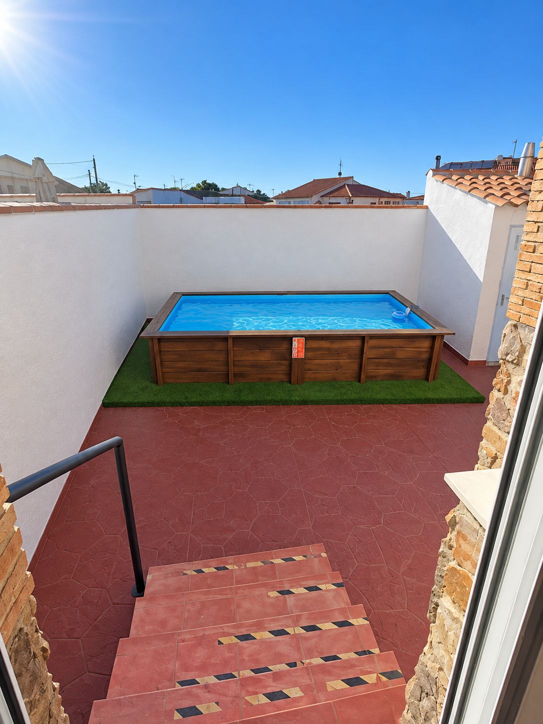 A swimming pool with a wooden deck, a red tiled floor, and a small grassy area.