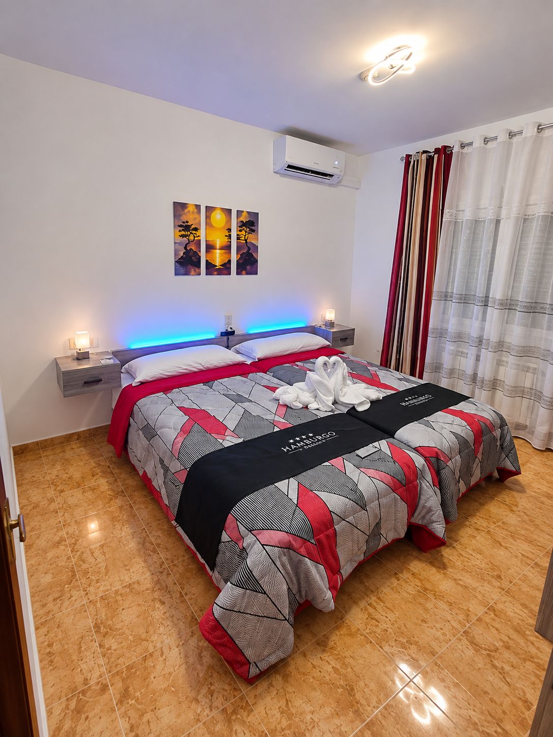 A hotel room with a bed, nightstands, an air conditioning unit, a wall-mounted artwork, and a window with curtains.
