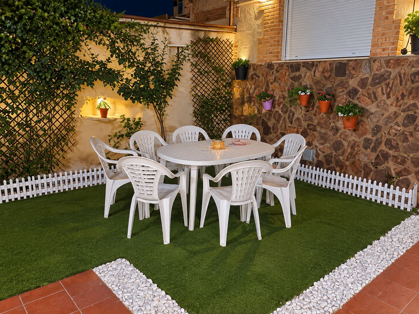 A patio with a round table and white chairs, surrounded by a white picket fence and a stone wall.