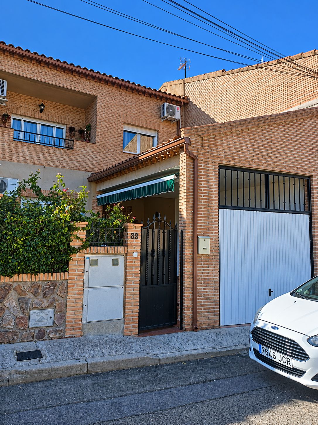 Una casa de ladrillo de dos pisos con una puerta de garaje blanca, una reja metálica y un automóvil blanco estacionado en la parte delantera.