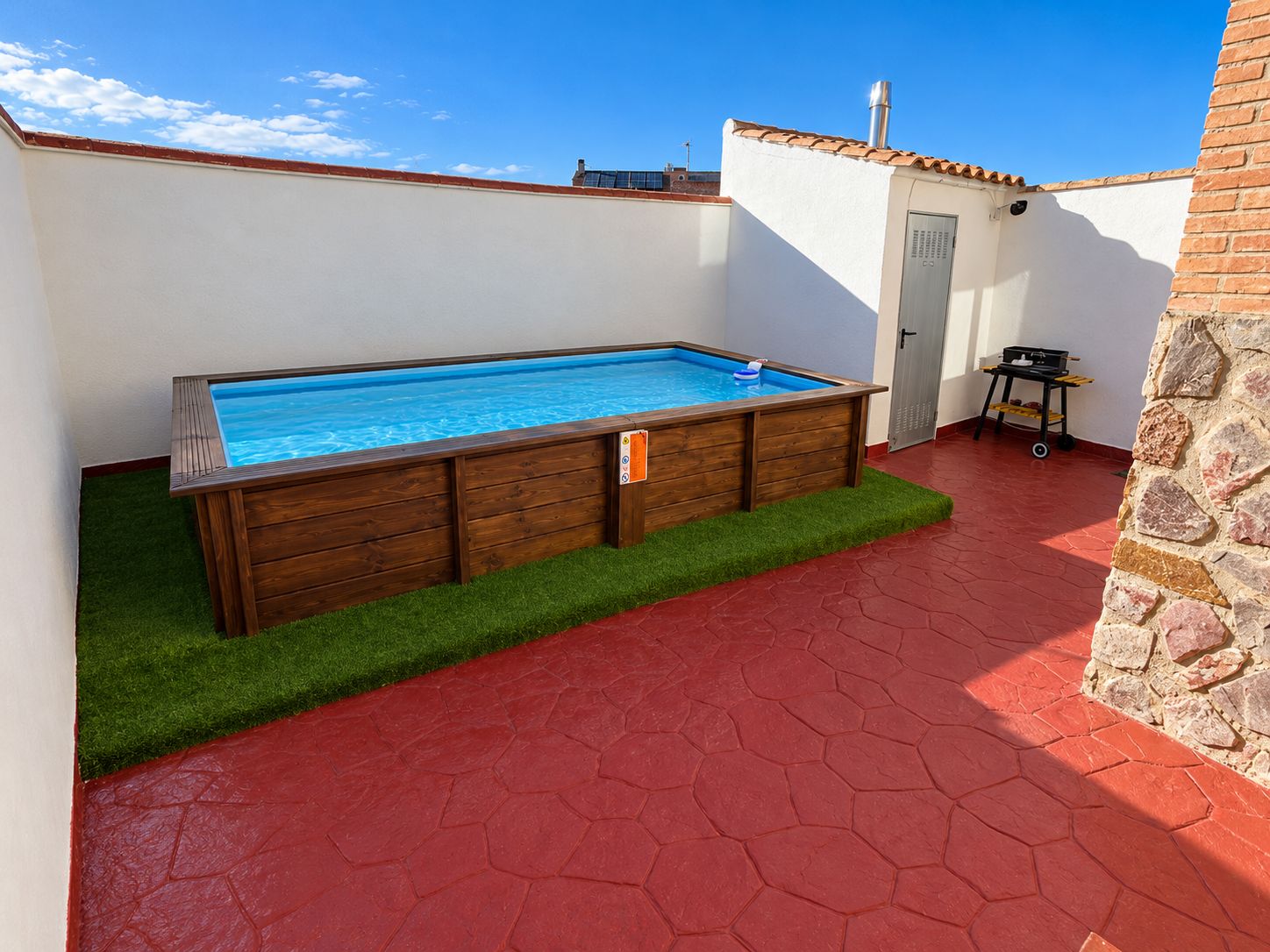 Una piscina con cubierta de madera, rodeada por un patio de baldosas rojas y un muro de piedra.