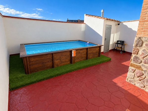 A swimming pool with a wooden deck, surrounded by a red tiled patio and a stone wall.