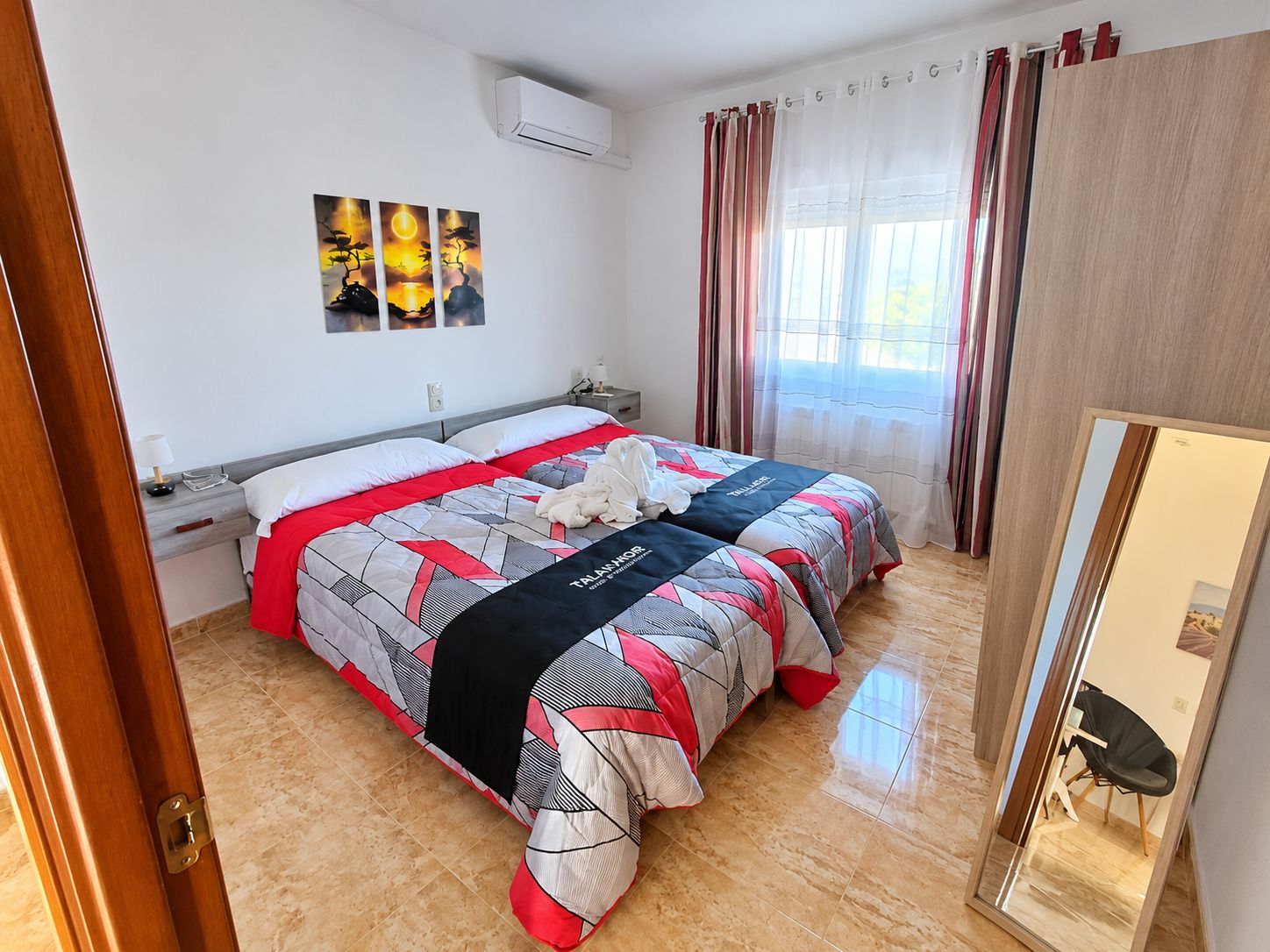 Una habitación de hotel con cama, mesita de noche, lámpara, aire acondicionado instalado en la pared y ventana con cortinas.
