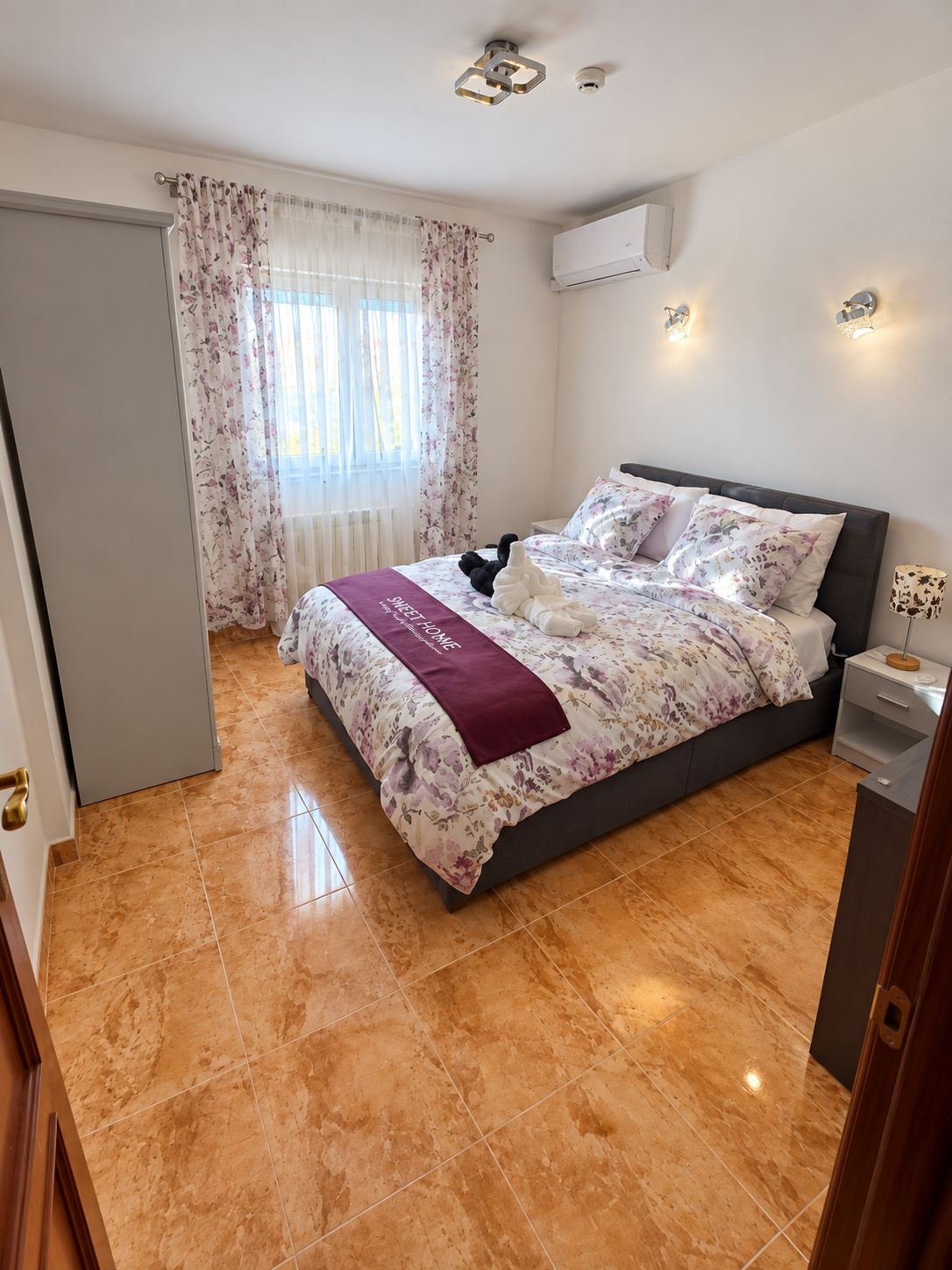 Una habitación de hotel con cama, mesita de noche, lámpara, ventana con cortinas y aire acondicionado.