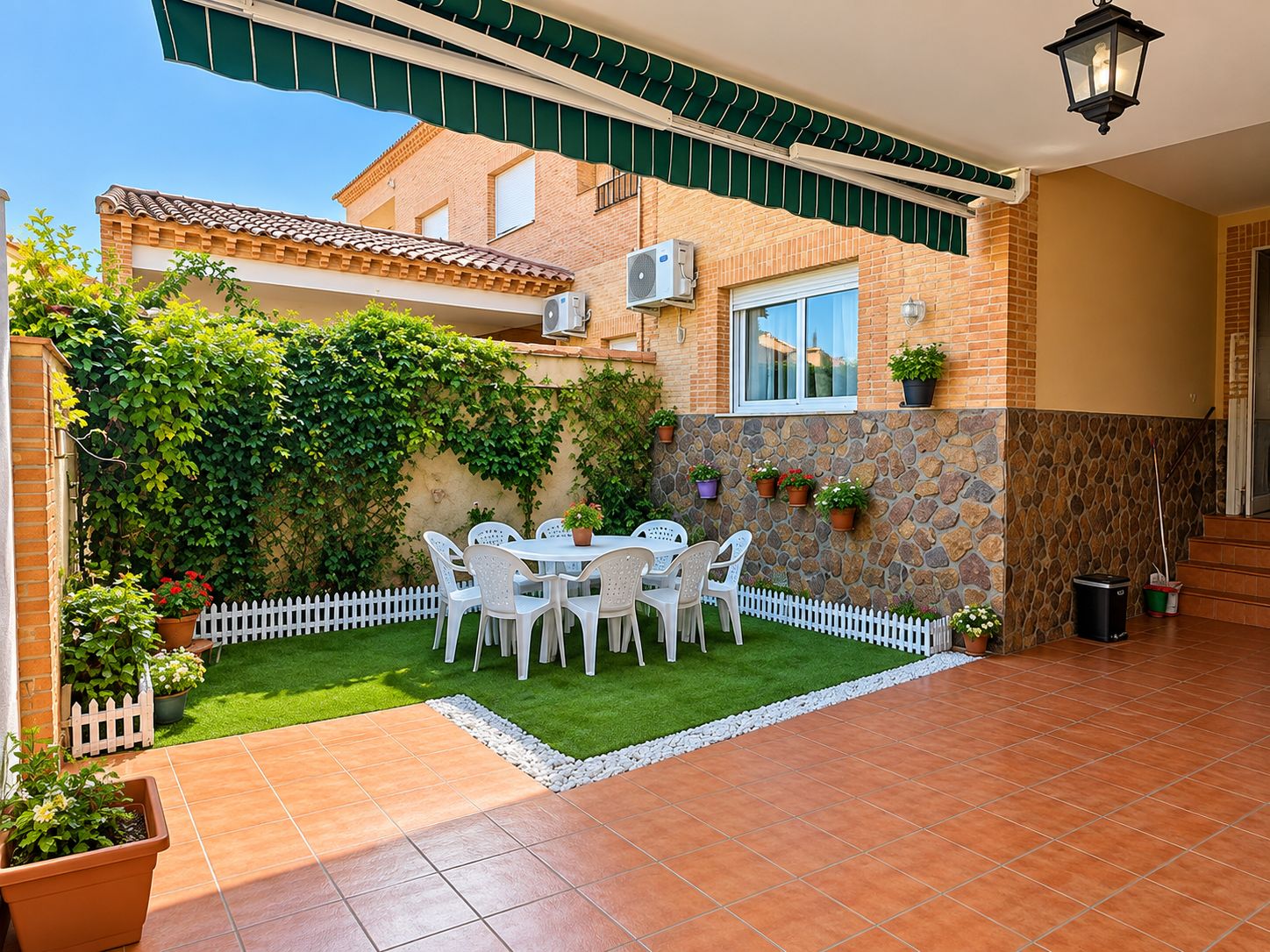 Un patio con una mesa y sillas blancas, rodeado por una cerca de piquetes blancos y un muro de piedra con plantas en macetas.