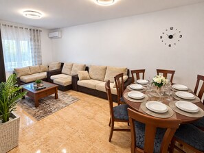 Een woonkamer met een hoekbank, een salontafel, een eettafel gedekt voor vier personen en een wandklok.
