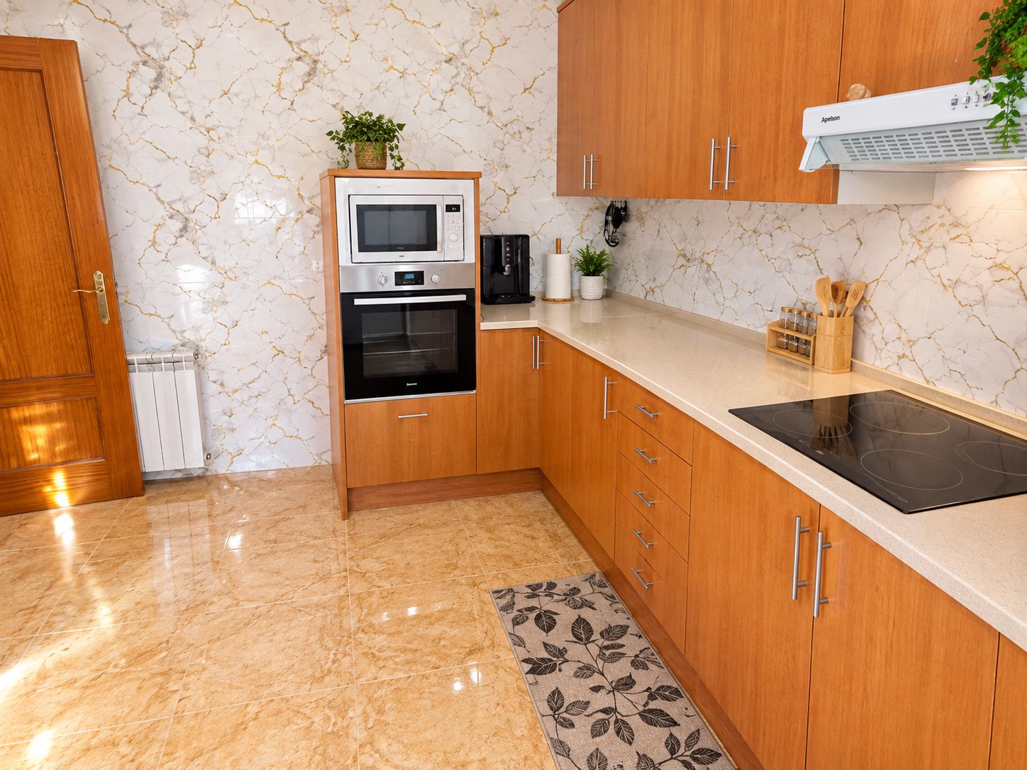 Una cocina con horno empotrado, microondas y estufa, gabinetes de madera, salpicadero de mármol y una alfombra pequeña.