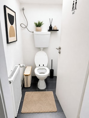 Un baño pequeño y moderno con inodoro blanco, un radiador, un felpudo beige, una planta en maceta y un estante negro con un florero y difusor de cañas.